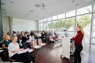 Eine Frau im roten Pullover spricht in einem großen, hellen Konferenzraum vor etwa 50 Personen.